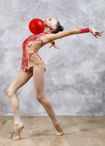 A young rhythmic gymnast in a photo pose wearing a leotard with red and purple colors, decorated with sequins and rhinestones, and a ball attached to her head.